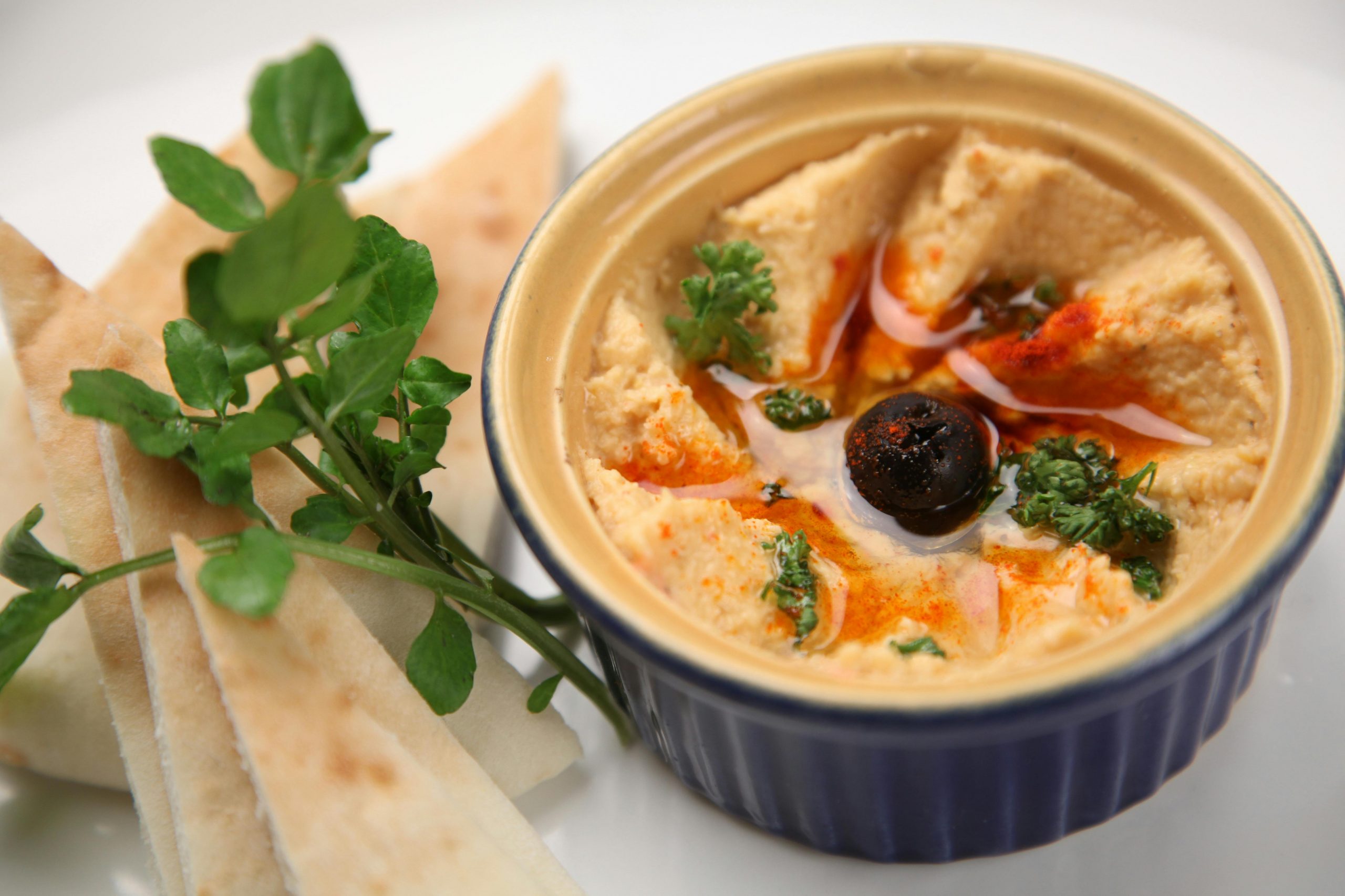 A close-up of creamy homemade hummus with pita slices, garnished with olive and herbs.