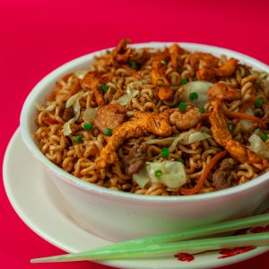 Bowl of spicy chicken noodles garnished with vegetables against a vibrant red background.