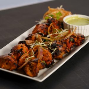 A flavorful chicken tikka dish served with a side of green chutney and garnishes on a white platter.