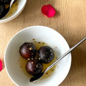 Close-up of gulab jamun in a bowl with rose petals on a wooden table, showcasing Indian dessert charm.