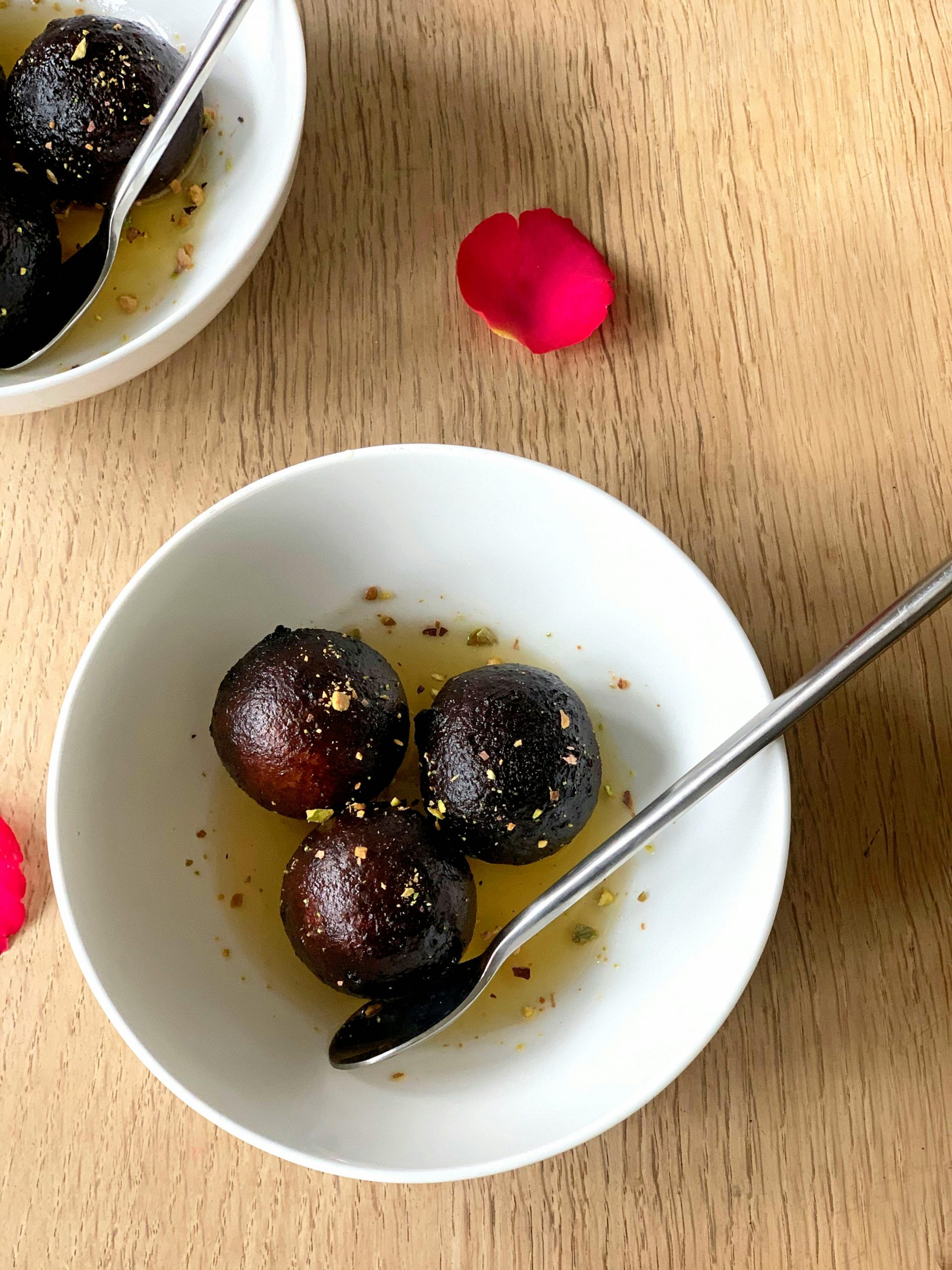 Close-up of gulab jamun in a bowl with rose petals on a wooden table, showcasing Indian dessert charm.